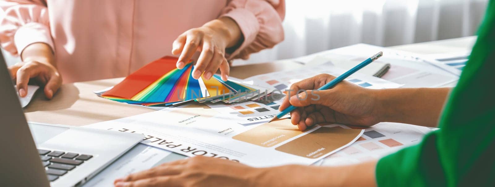 Cropped imaged of professional interior designers choose appropriate color from color palette with house model and architectural equipment scatter around. Closeup. Focus on hand. Variegated.