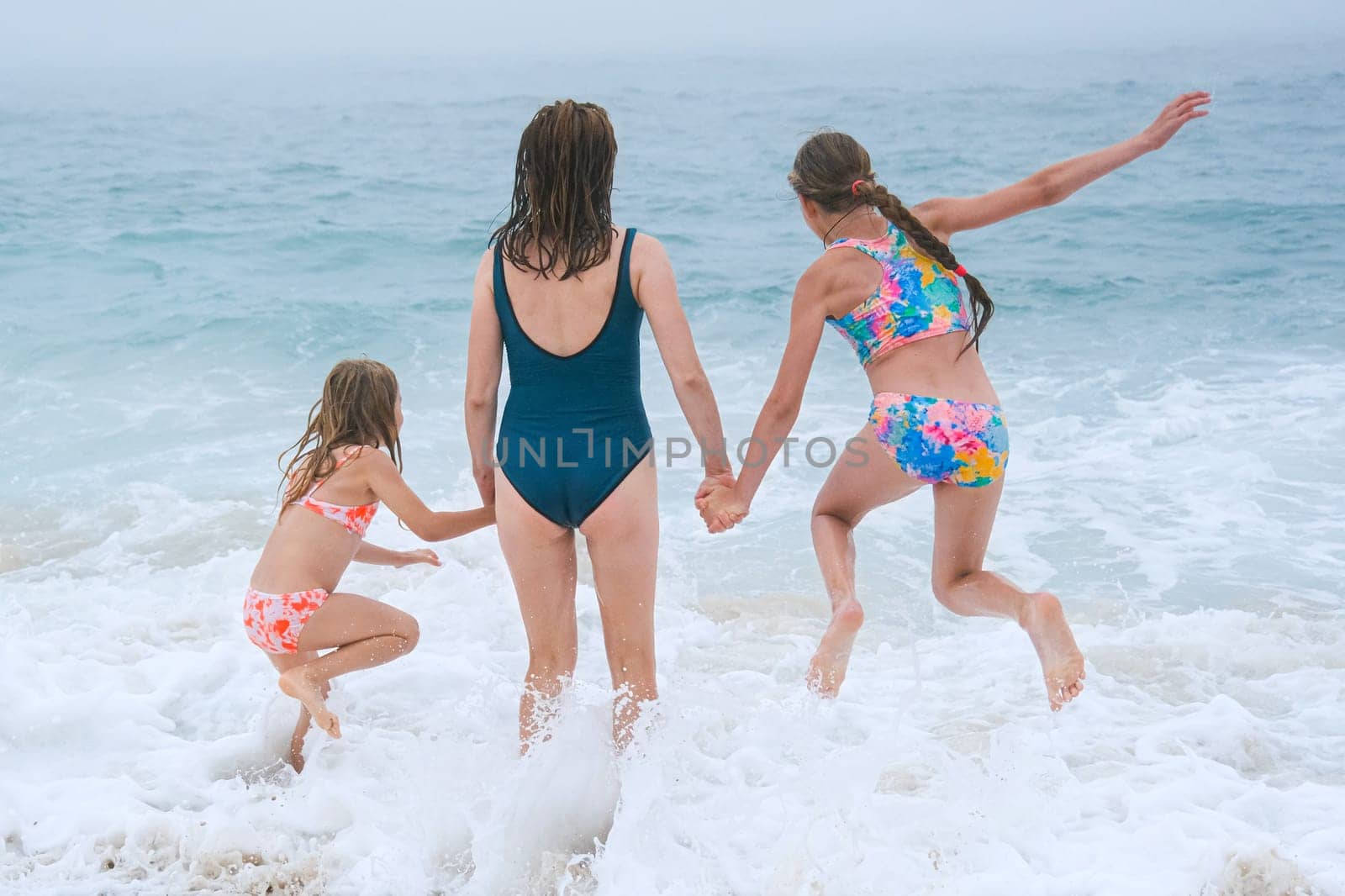 Mother and children playing on the ocean beach. Family enjoying the ocean. Mother holds girls's hands and they all look at the ocean together.