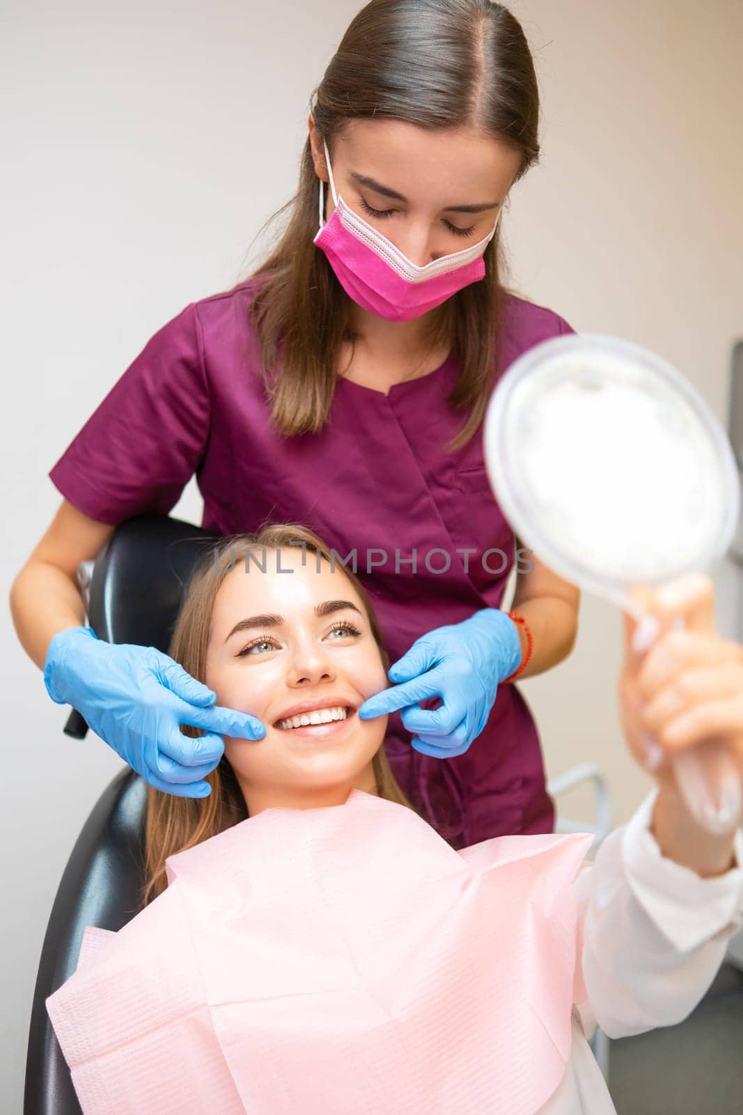 Young patient is looking in the mirror and admires her perfect smile after dental treatment by vladimka