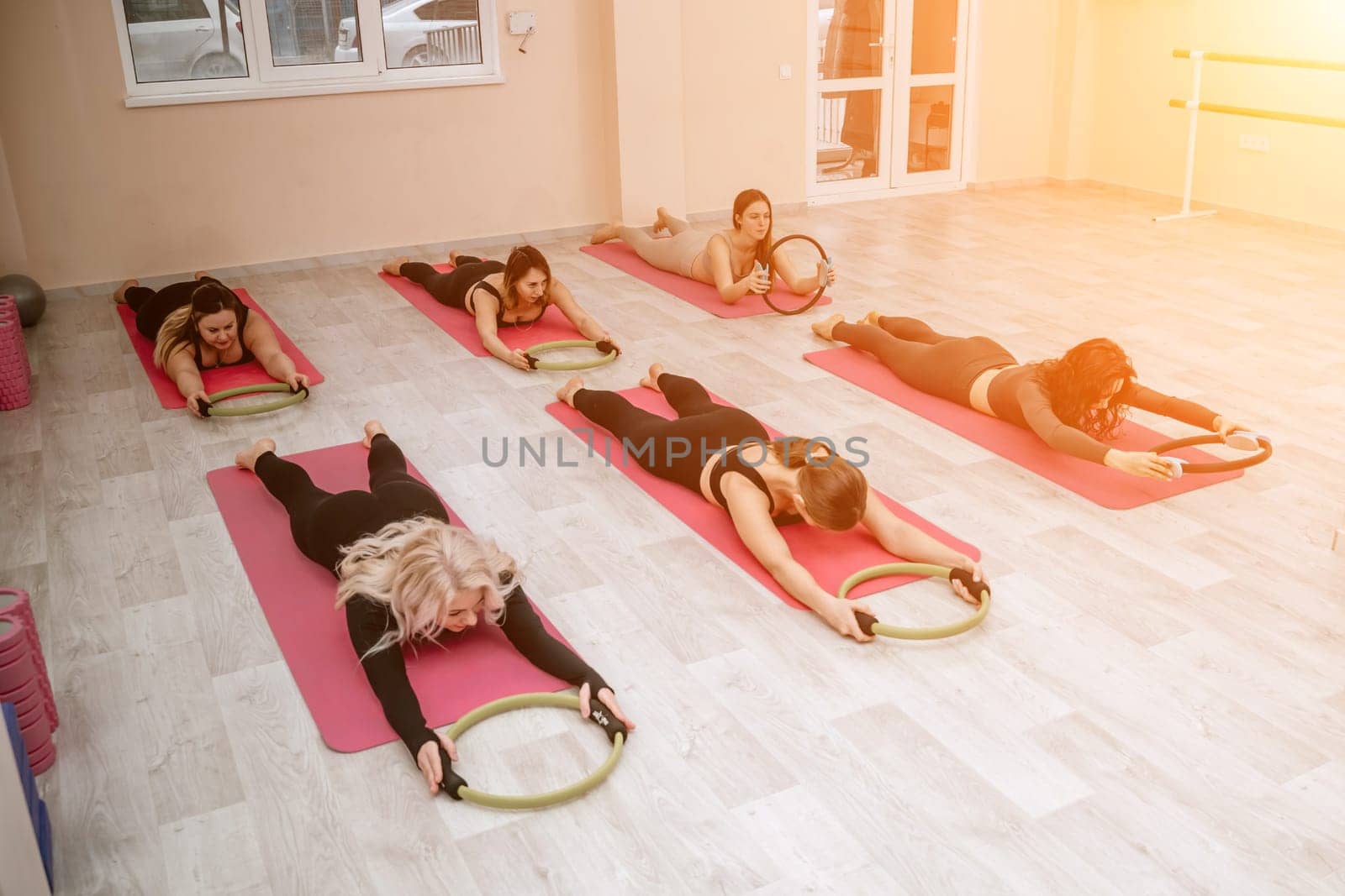 A group of six athletic women doing pilates or yoga on pink mats in front of a window in a beige loft studio interior. Teamwork, good mood and healthy lifestyle concept. by Matiunina