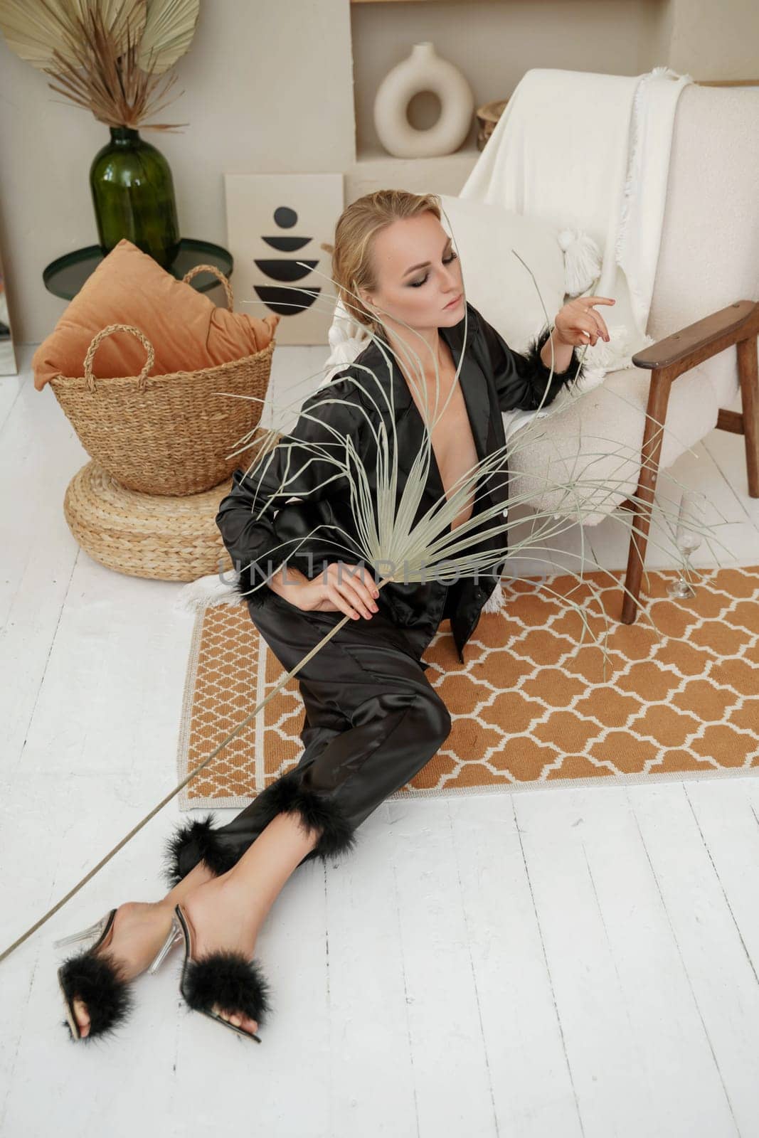 Woman glamorous pajamas. A beautiful blonde in black pajamas is sitting relaxed in a chair in black pajamas with feathers. Beige stones are all over the interior