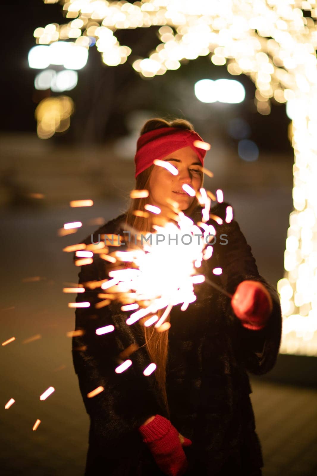 Woman holding sparkler night while celebrating Christmas outside. Dressed in a fur coat and a red headband. Blurred christmas decorations in the background. Selective focus.