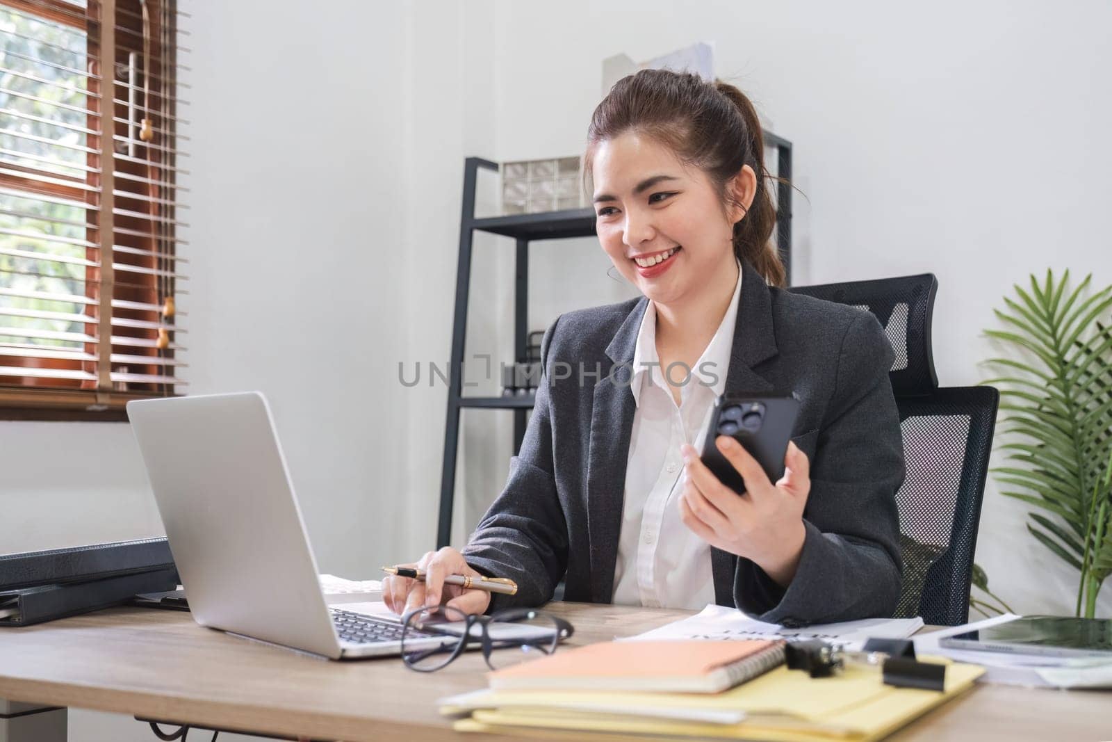 Businesswoman talking on business phone and using laptop to do finance, mathematics on wooden table in office and business background, tax, accounting, statistics and analytical research concepts. by wichayada