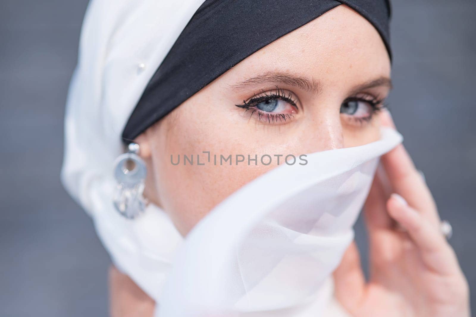 Portrait of a young blue-eyed woman in a hijab against a gray brick wall. Muslim woman with white scarf covering her face. by mrwed54