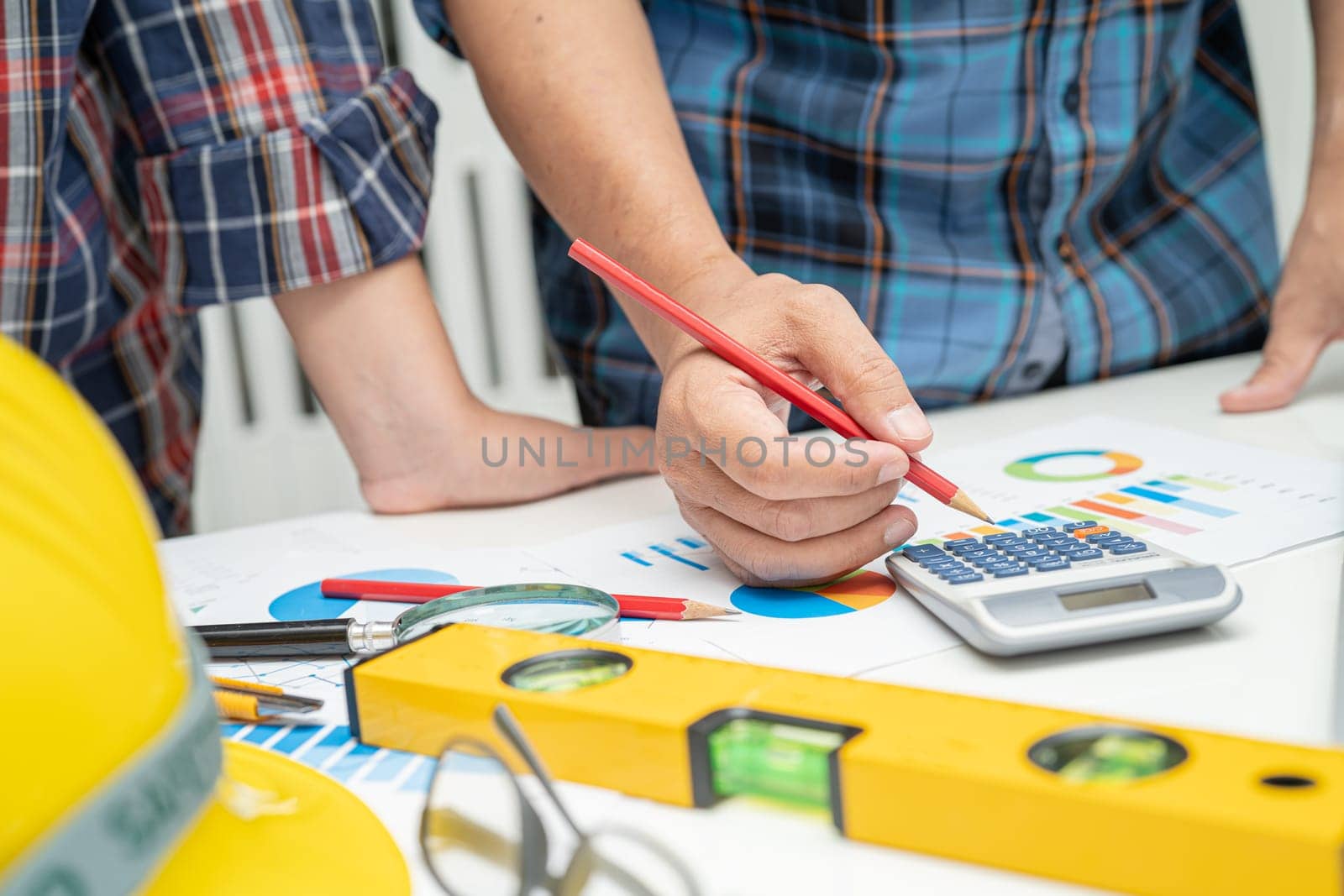 Asian engineer and partner team learn and talk with house plan project blueprint, graph and yellow helmet and engineering construction tools on table work. by pamai