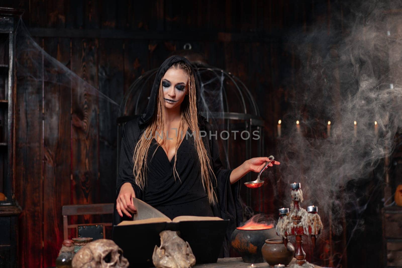 Halloween, witch use magic book and cauldron prepare poison or love potion. Black magic occult female wizard in dark room cage spider web human skull. Dressed black cloak with dreadlocks
