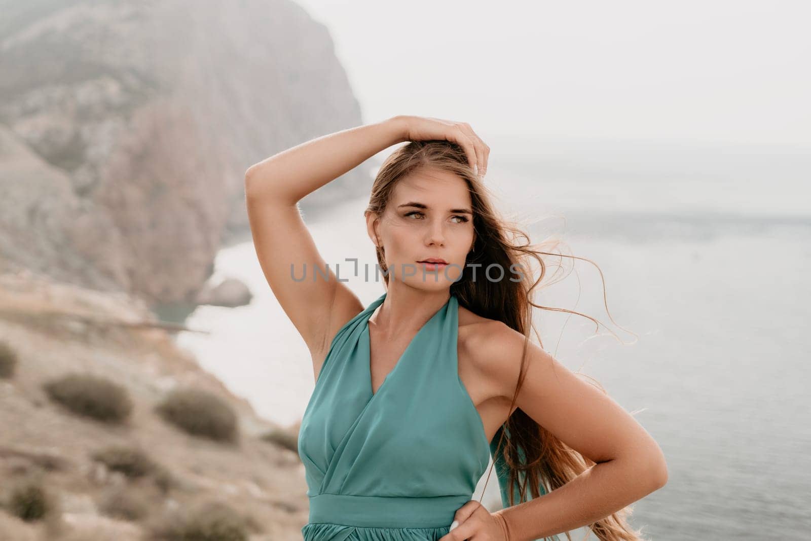 Side view a Young beautiful sensual woman in a mint long dress posing on a volcanic rock high above the sea during sunset. Girl on the nature on overcast sky background. Fashion photo