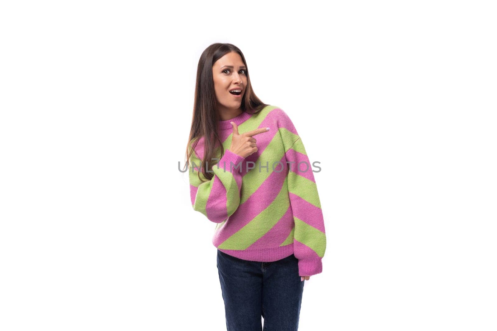 stylish cheerful young brunette woman in a striped pink sweater points her finger to the side on a white background with copy space.