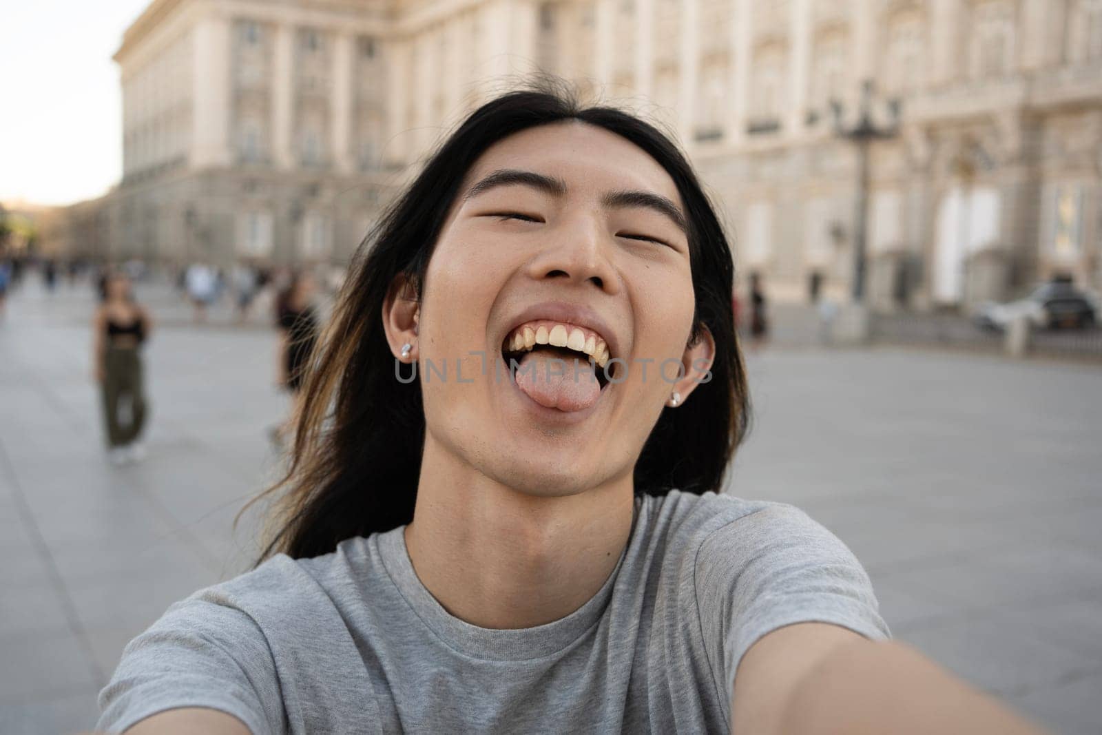 Asian young guy showing his tongue while taking selfie photo in Madrid city by papatonic