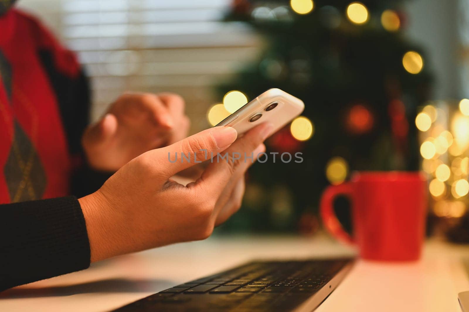 Cropped shot of young woman using mobile phone near Christmas tree at cozy home by prathanchorruangsak