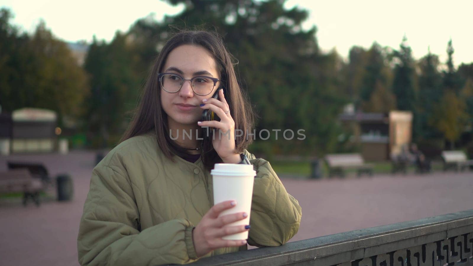 A young woman speaks on the phone with a glass of coffee in her hand. A girl in glasses with a phone near the fence on the embankment with a hot drink in a glass. 4k