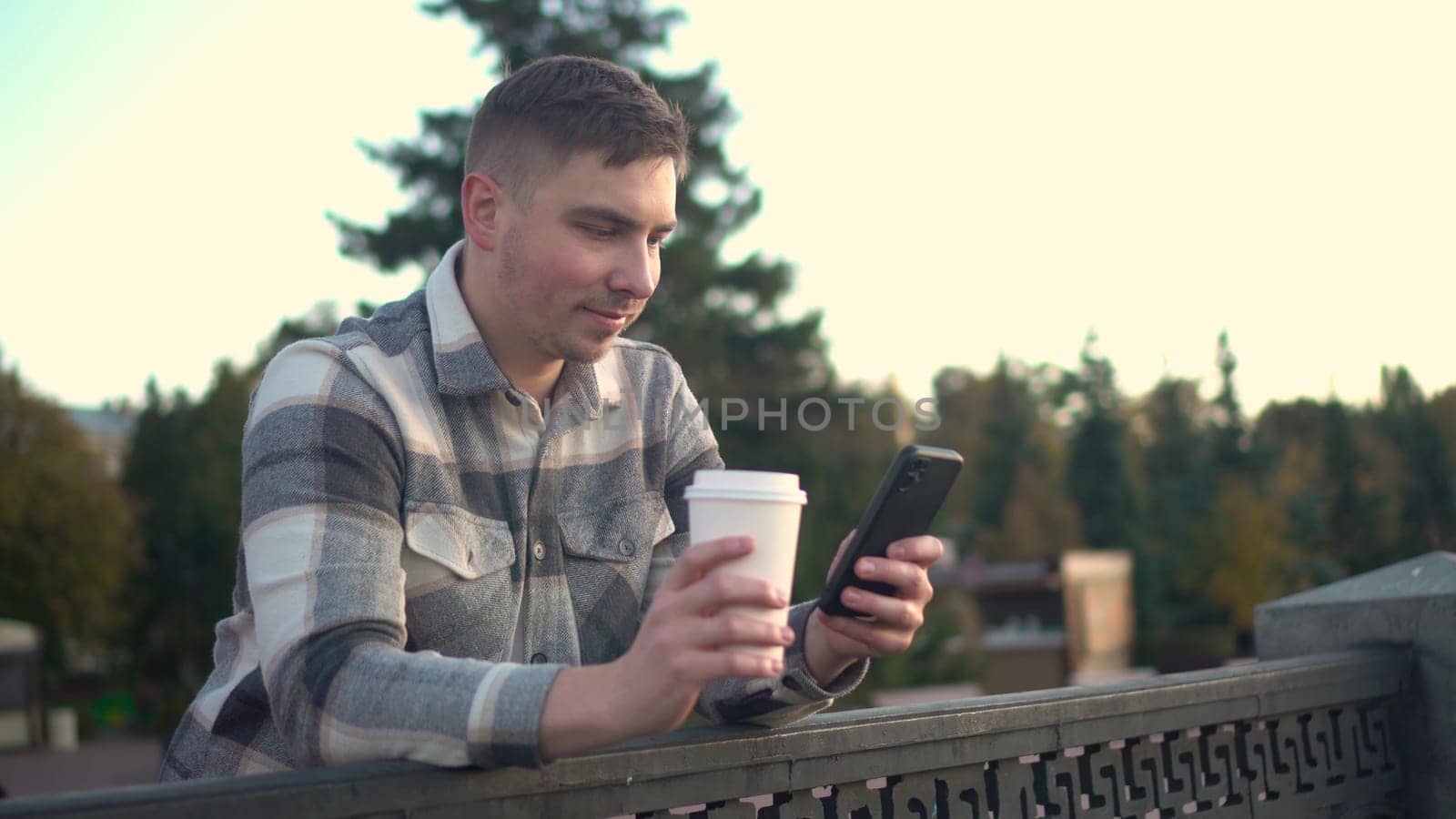 A young man is chatting on a smartphone with a glass of coffee in his hand. A man with a phone near the fence on the embankment with a hot drink in a glass. 4k