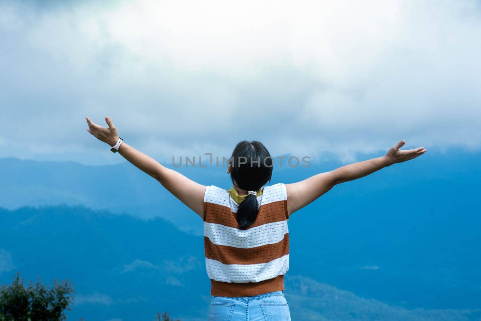 Female traveler enjoying the beauty of nature on the mountain looking at the lush trees in the tropical forest. Hipster woman raises her arms and enjoys the mountaintop view. by TEERASAK