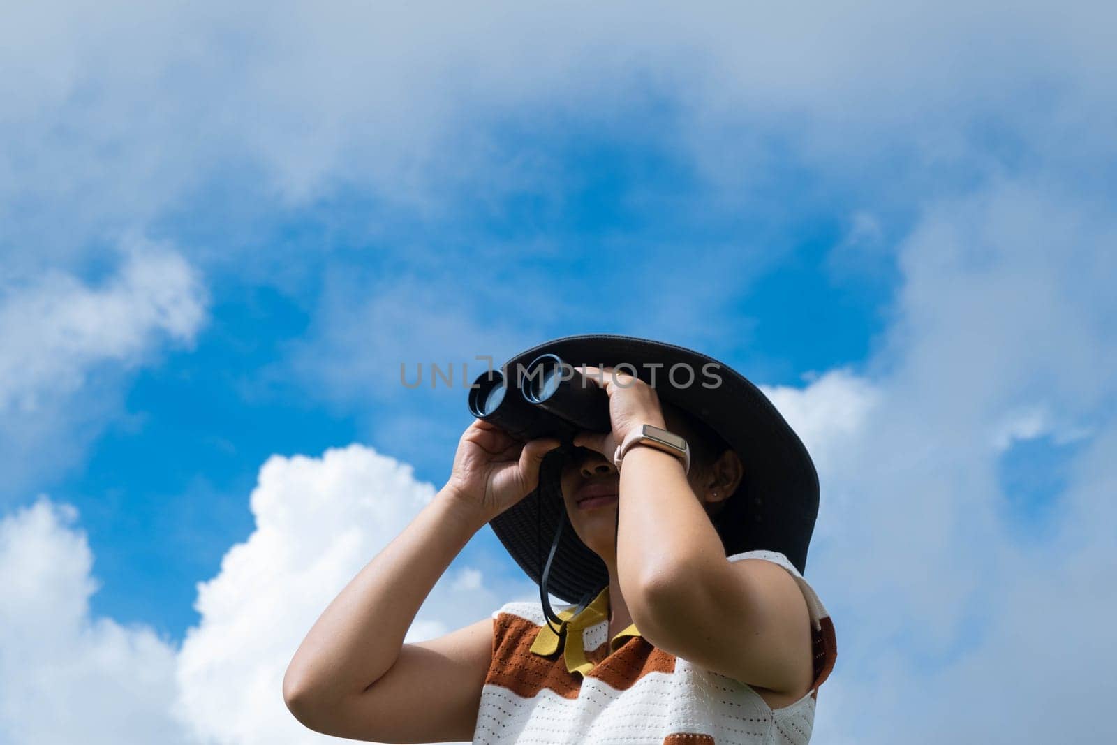 Young woman with binoculars on the mountain on a sunny day. Woman using binoculars when going hiking. Hiking woman uses binoculars to travel and has a happy smile.