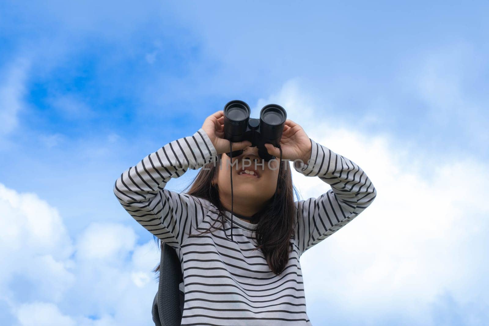 Cute little girl with binoculars on the mountain on a sunny day. Young girl uses binoculars when going hiking. Active young girl uses binoculars on a trip and smiles happily.