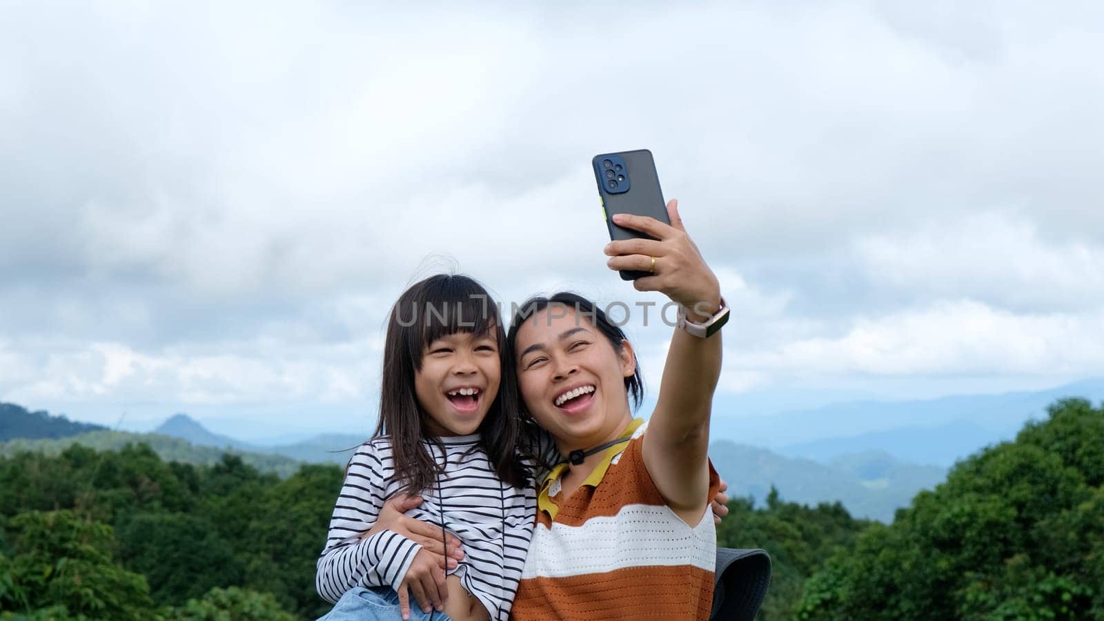 Cute little Asian girl and mother take selfie together with beautiful view of green mountains in summer. A girl traveling with mother. A woman is photographed with her daughter, Camping in the mountains with children. by TEERASAK