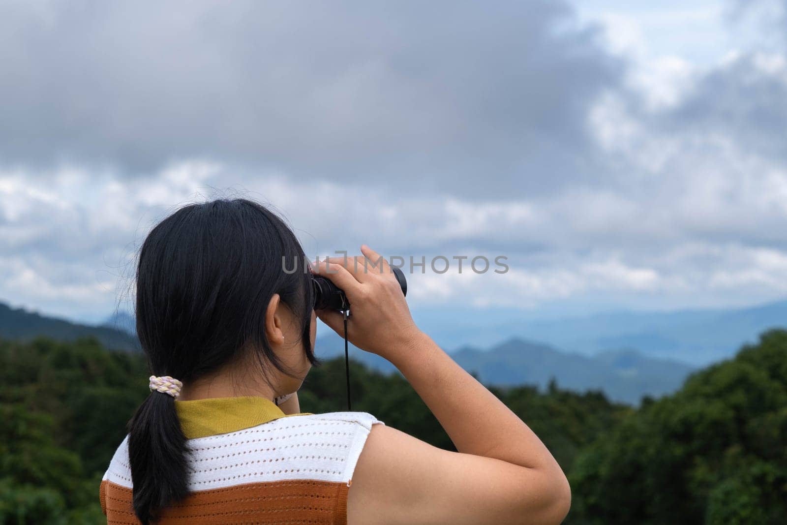 Young woman with binoculars on the mountain on a sunny day. Woman using binoculars when going hiking. Hiking woman uses binoculars to travel and has a happy smile. by TEERASAK