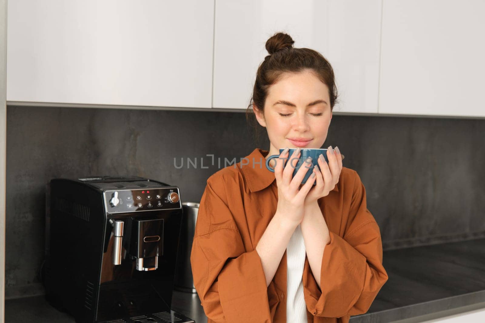 Portrait of beautiful young woman, enjoying delicious aroma of freshly made coffee, holding mug, standing near machine in the kitchen, drinking from mug.