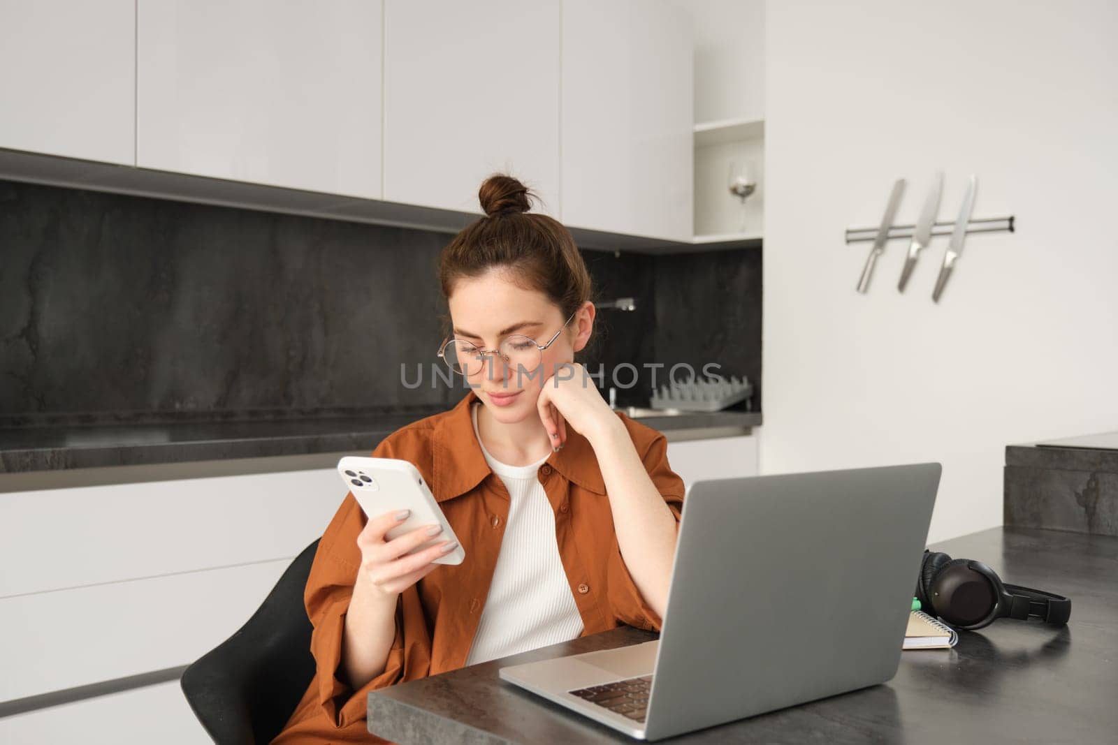 Portrait of young self-employed woman working on start up from home, freelancer programmer with computer, doing her task on computer, checking her mobile phone, reading message on smartphone.