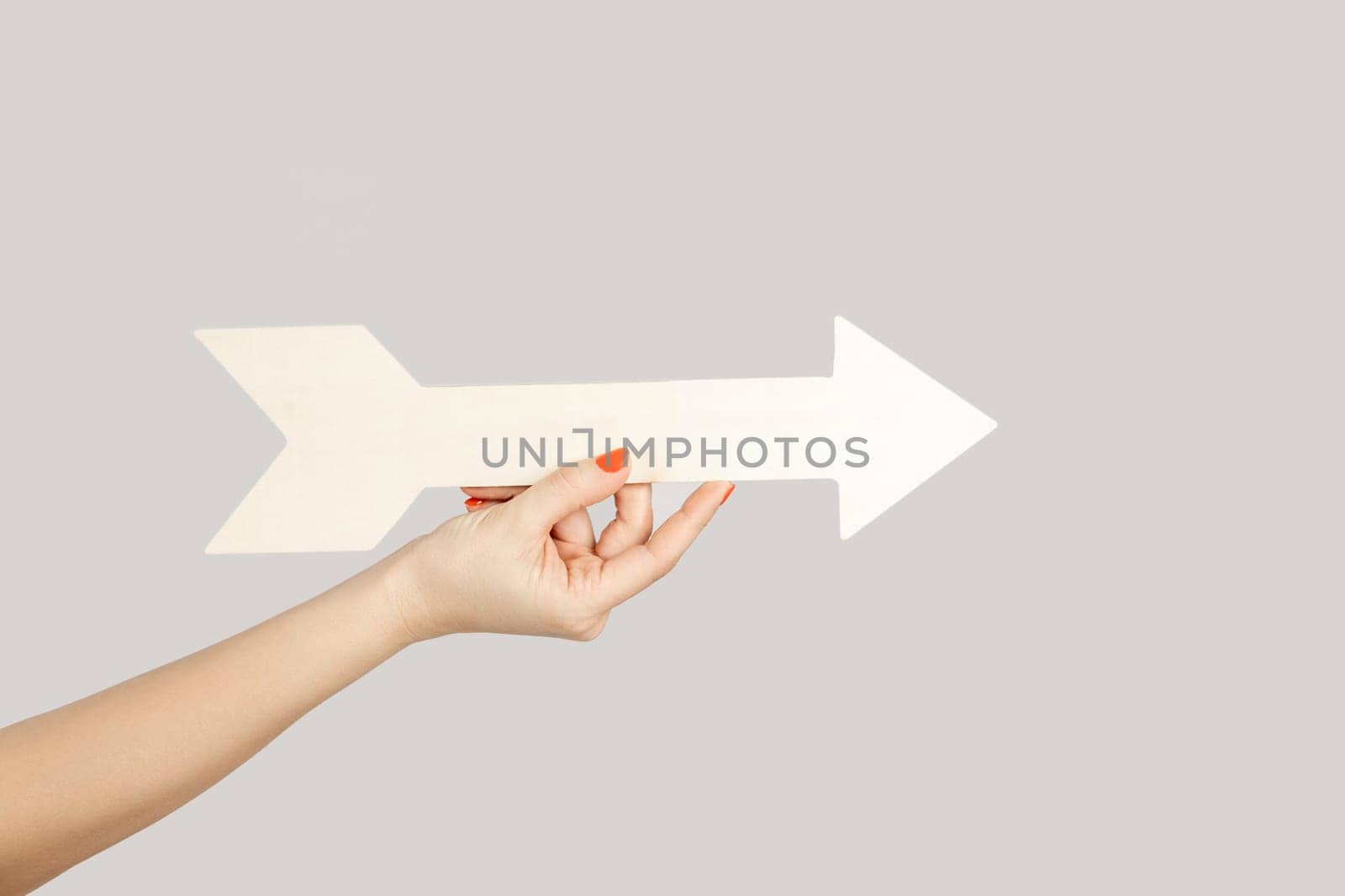 Closeup of woman hand holding white arrow pointing away, showing direction aside. Indoor studio shot isolated on gray background.