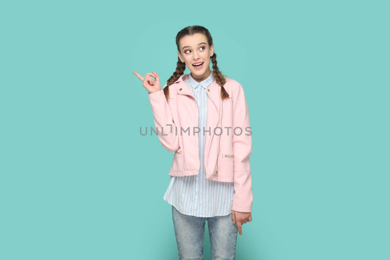 Portrait of excited smiling positive teenager girl with braids wearing pink jacket pointing index finger away, showing copy space for advertisement. Indoor studio shot isolated on green background.