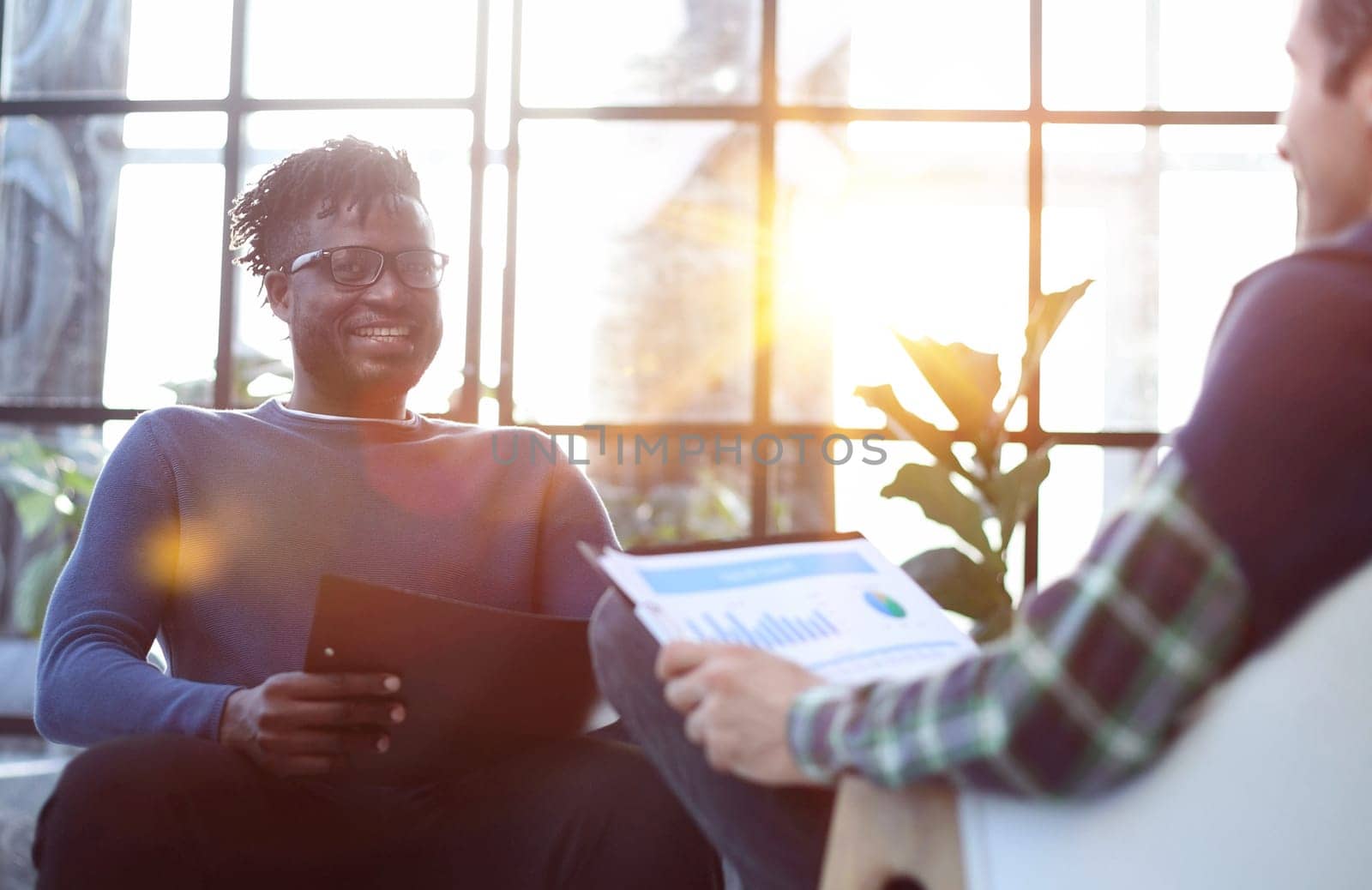 black man with a folder in his hands laughs while sitting on a chair