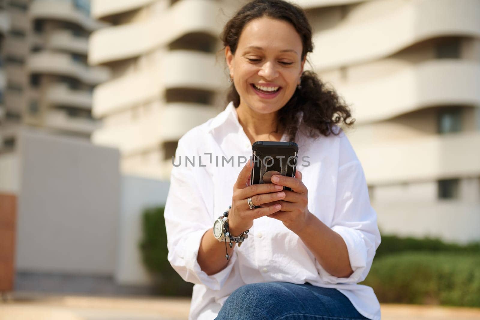 Multi ethnic brunette, beautiful smiling woman, business person, entrepreneur, student expressing positive emotions while using smartphone outdoors, sitting against cityscape background. Mobile app