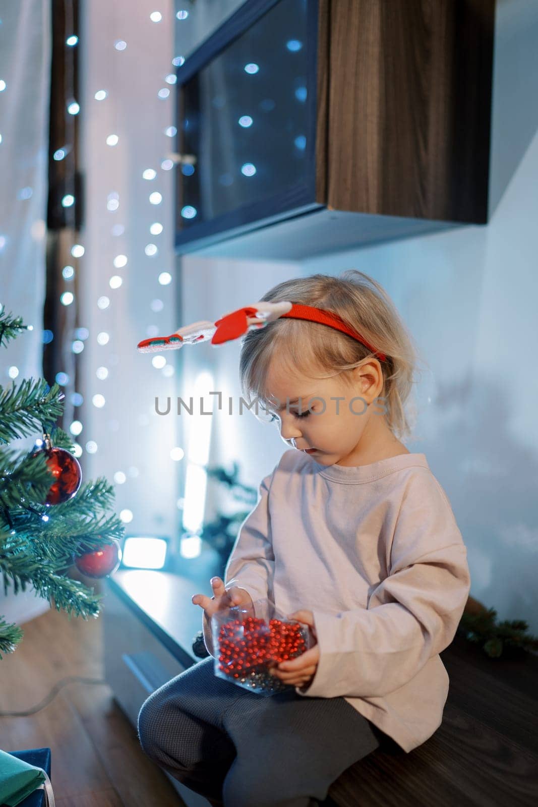 Little girl in a hoop examines red garlands-beads in a box while sitting on a low locker near the Christmas tree. High quality photo