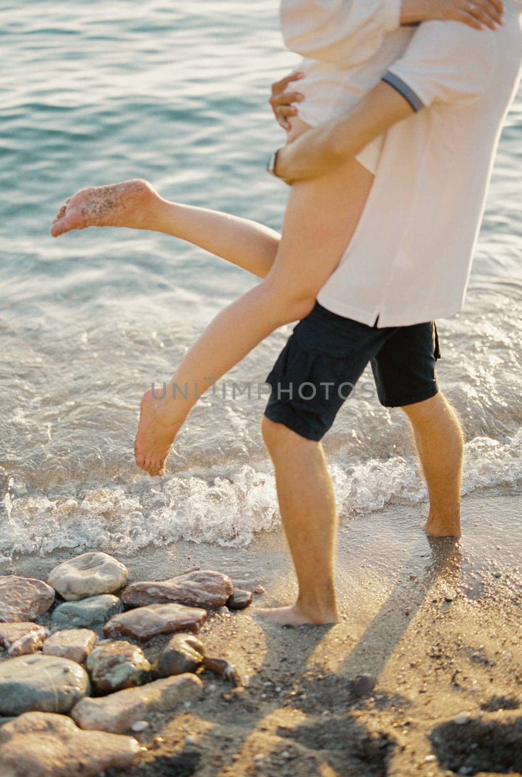 Man holds in his arms woman waving her legs while standing on the seashore. Cropped. Faceless by Nadtochiy