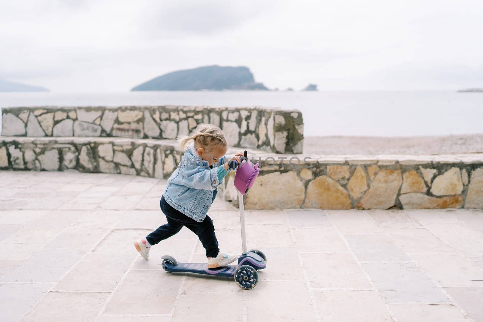 Little girl rides a scooter on a paved area near the sea looking down at her feet. High quality photo