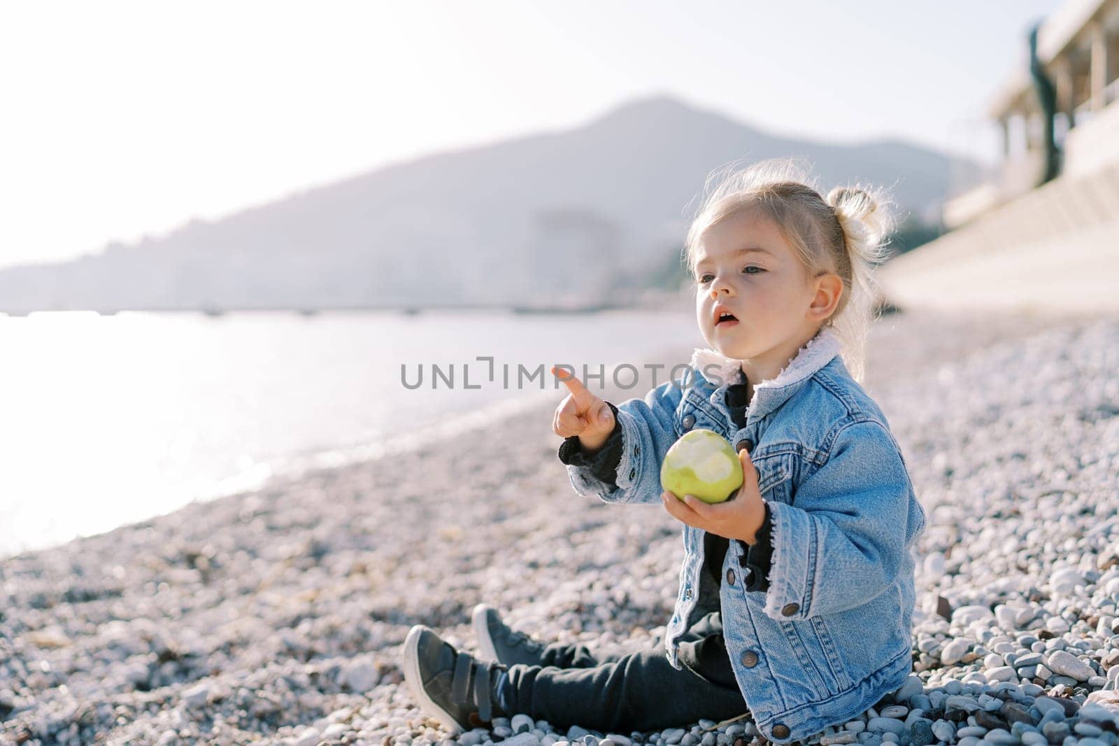 Little girl with an apple sits on a pebble beach and points to the sea. High quality photo