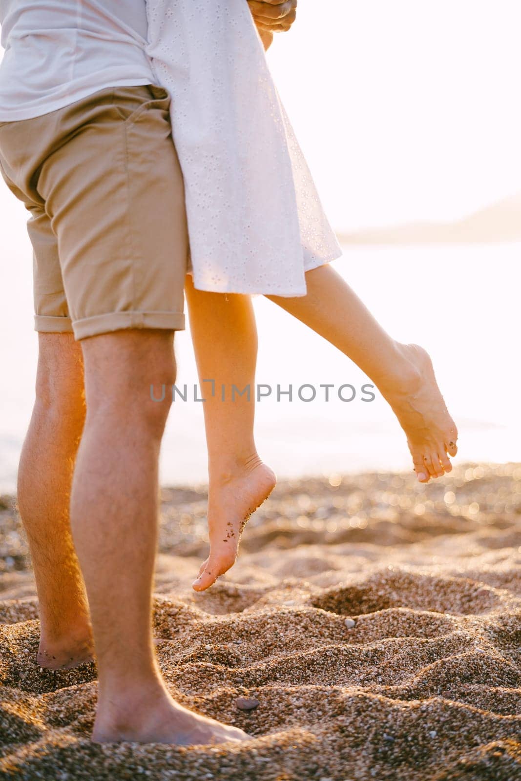 Man lifted up a woman dangling her bare feet while standing on the beach. Cropped. Faceless by Nadtochiy