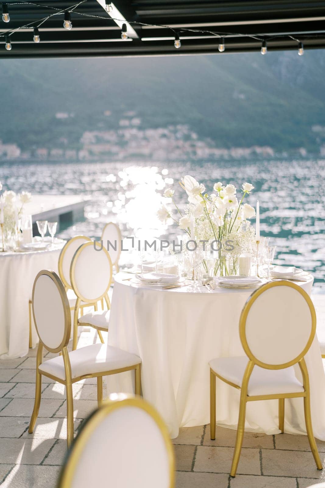 White chairs stand around round festive tables with bouquets of flowers under a cover on the pier. High quality photo