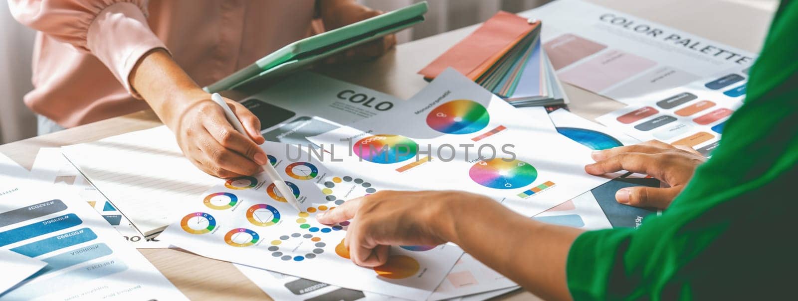 Skilled architect searching data from tablet while selecting an appropriate color from color wheel at table with color palette and document scatter around. Creative design concept. Variegated.