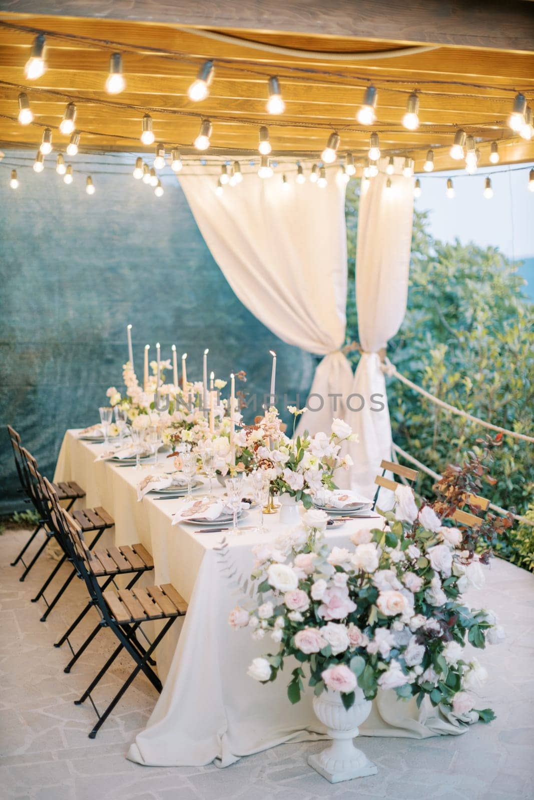 Long set table with bouquets of flowers and lit candles stands on a terrace with curtains by Nadtochiy