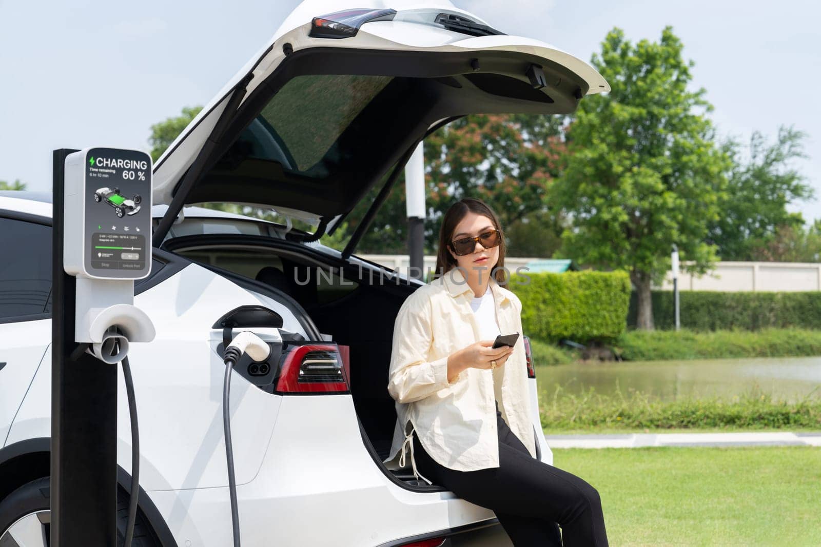 Young woman use smartphone to pay for electricity at public EV car charging station green city park. Modern environmental and sustainable urban lifestyle with EV vehicle. Expedient