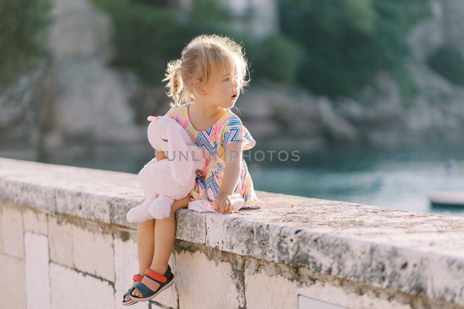 Little girl sits hugging a plush hare on a stone fence, looking back. High quality photo