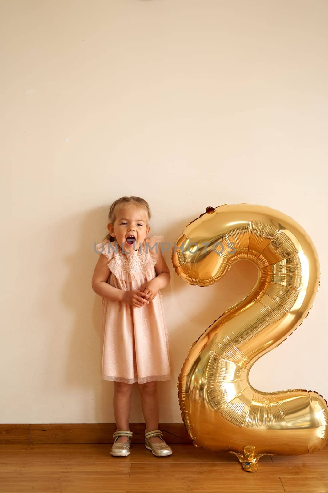 Little laughing girl stands against the wall near the inflatable golden number two. High quality photo
