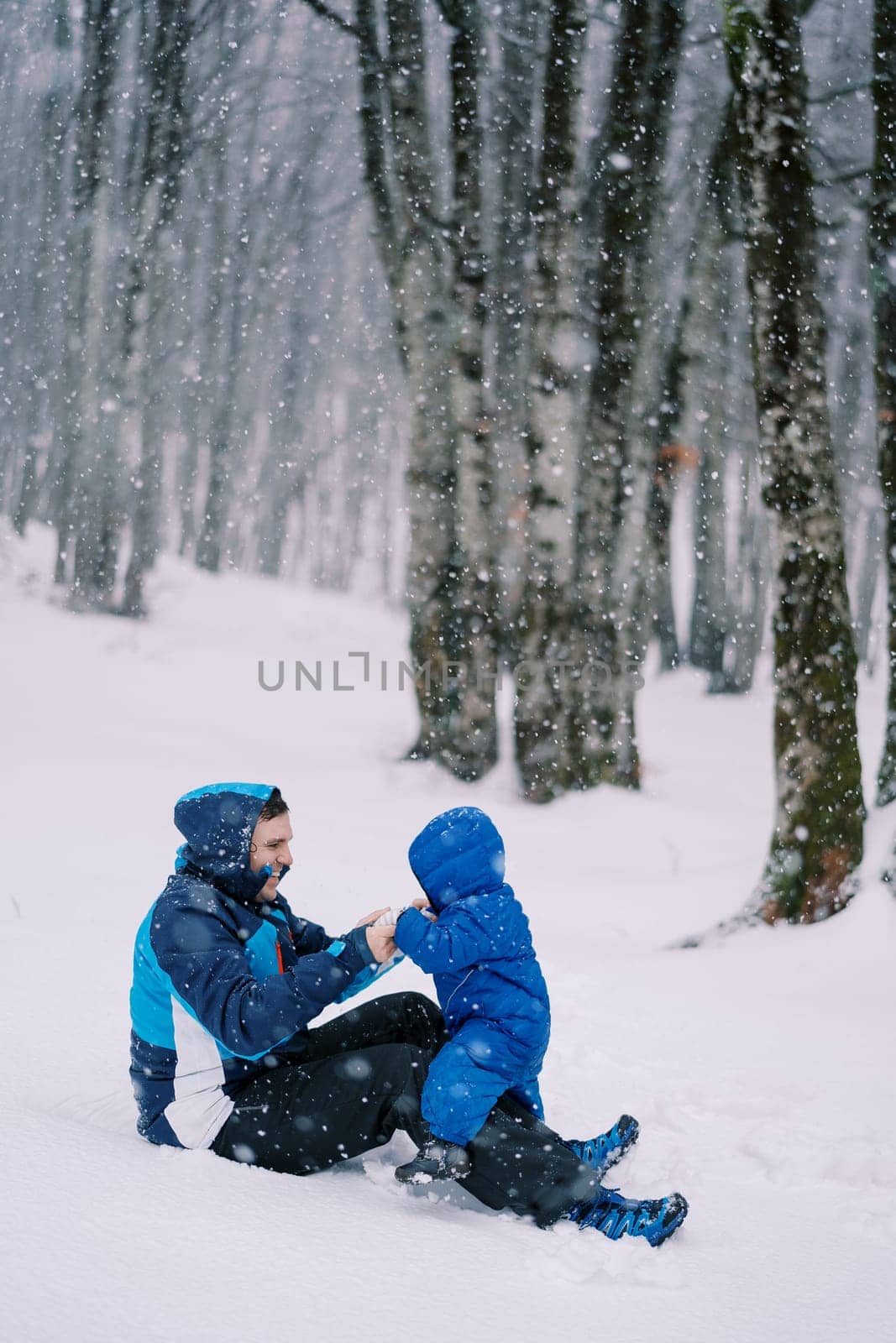 Laughing dad holds the hands of a small child sitting on his lap during a snowfall in the forest. High quality photo