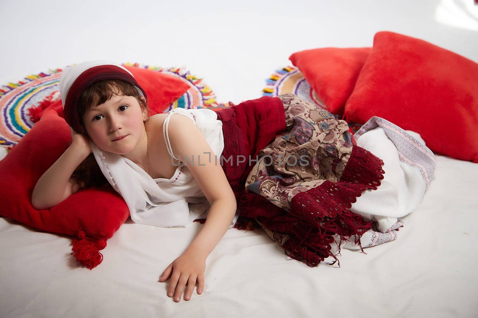 Portrait of Little girl in a stylized Tatar national costume having rest with a red pillow on a white background in the studio. Photo shoot of funny young teenager who is not a professional model