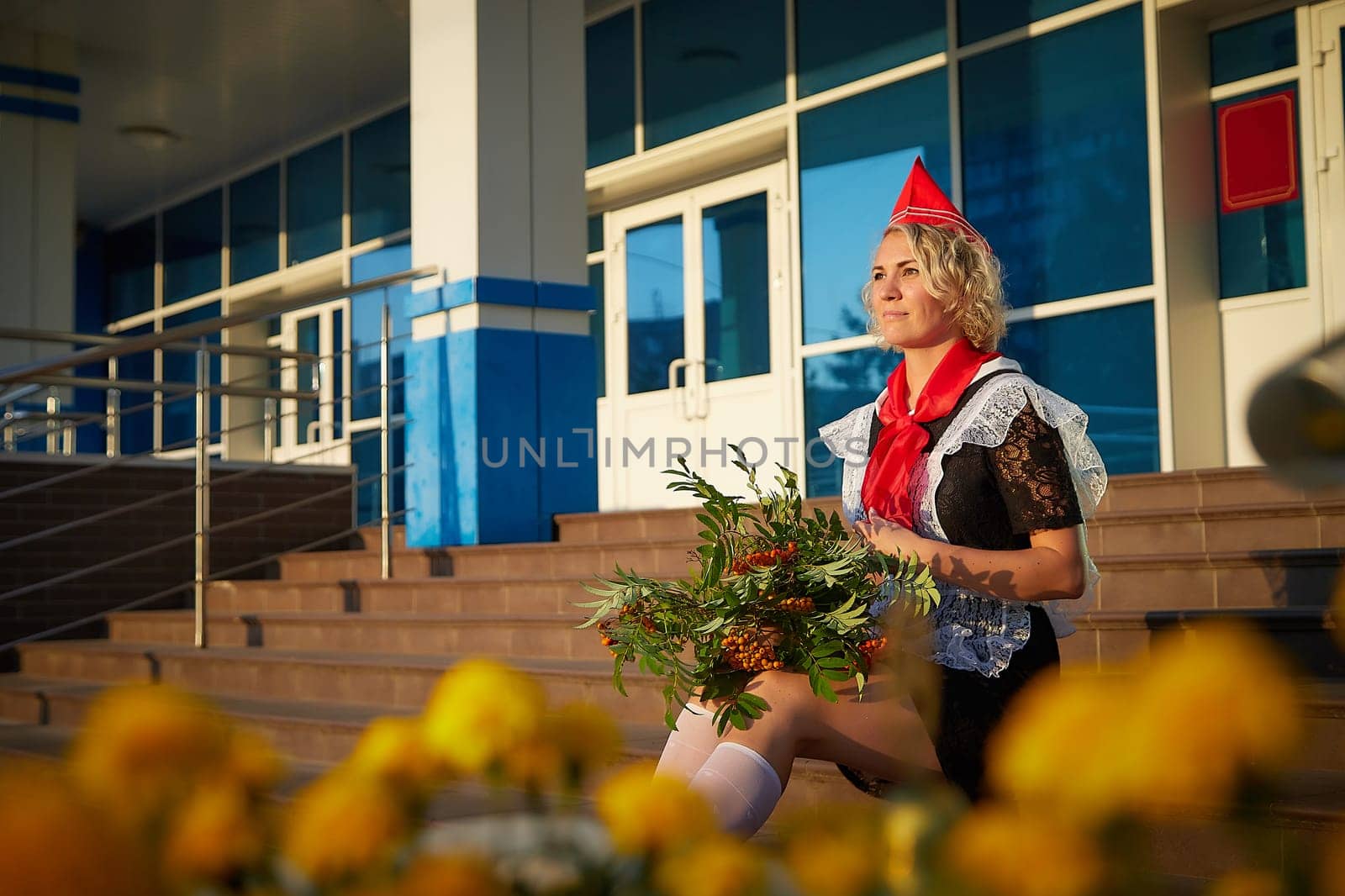 A Girl in black school uniform, white apron and red tie on steps of school with bouquet of flowers. Nostalgia photo shoot of teenager of female pioneer from USSR costume for September 1 or graduation