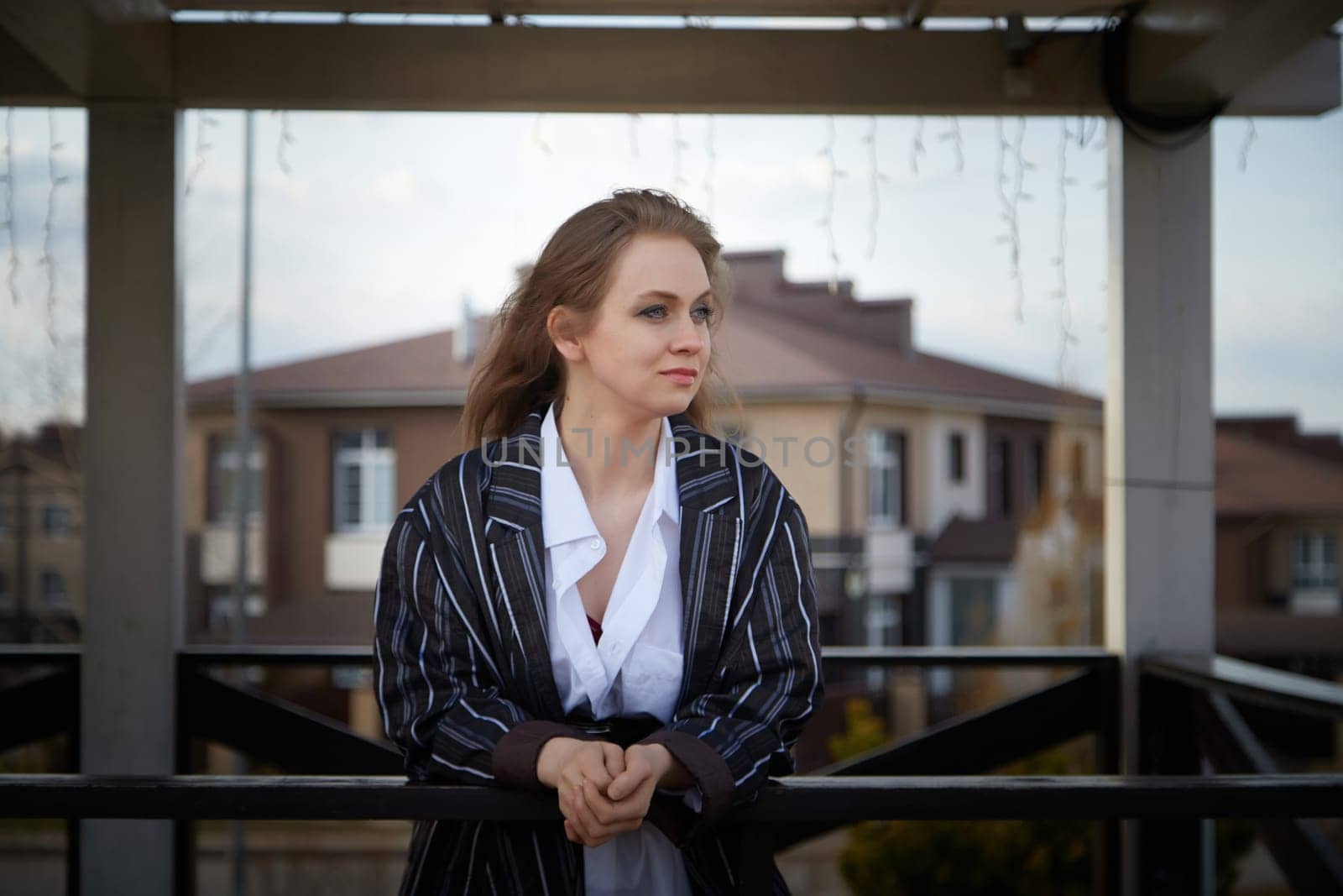 Beautiful girl with long hair in white shirt in open wooden pavillion in village or small town. Young slender woman in an autumn, spring or summer day