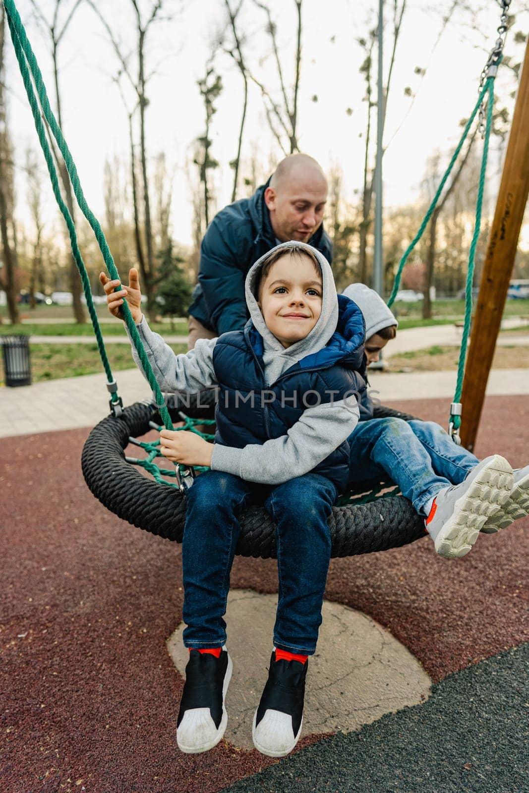 Father swinging his two little sons on a swing on playground by Fabrikasimf