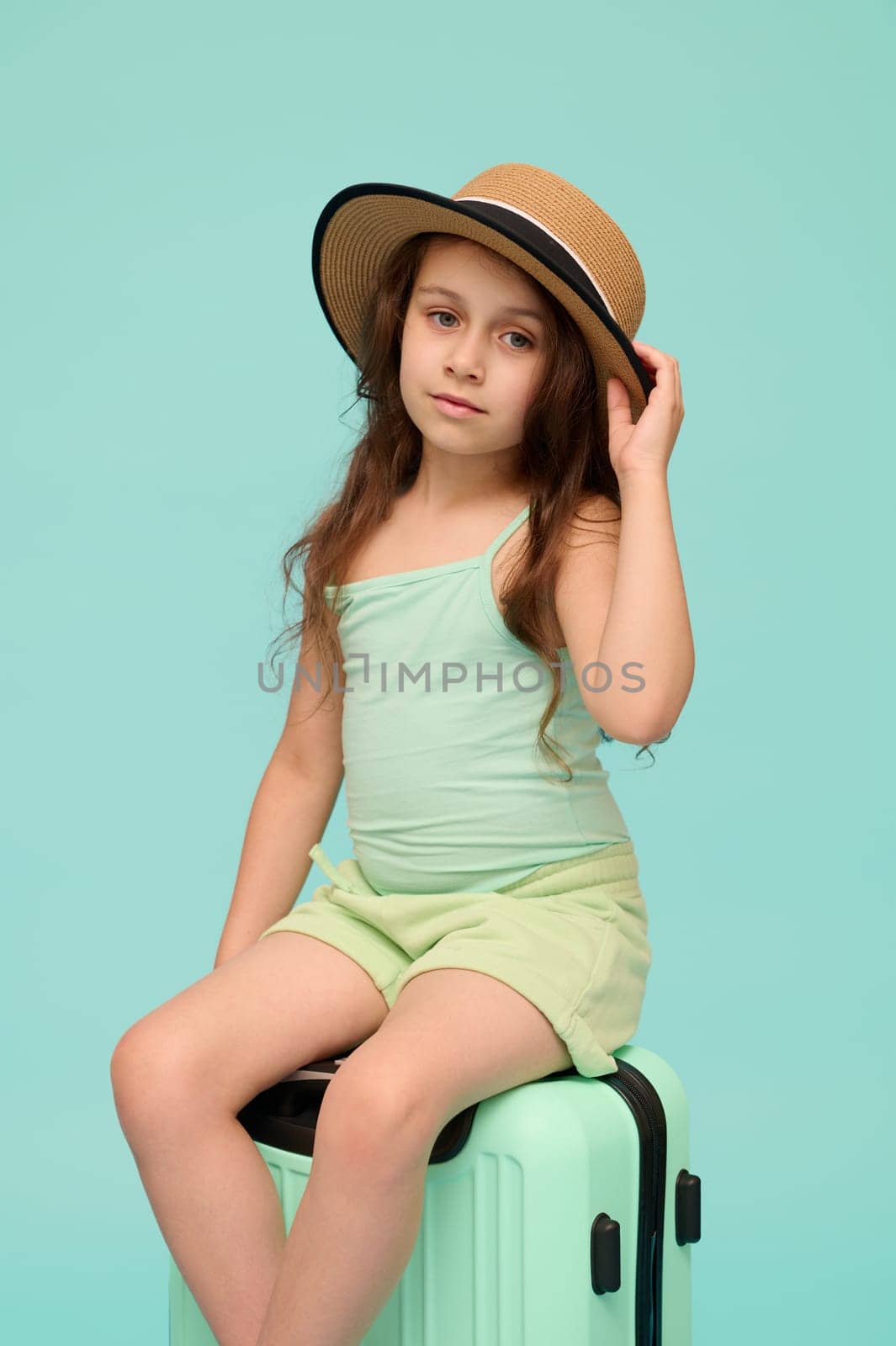 Vertical studio shot of a Caucasian little child girl 6 years old, dressed in summer clothes, smiles looking at camera, sitting on her suitcase, awaiting her flight, isolate over blue background