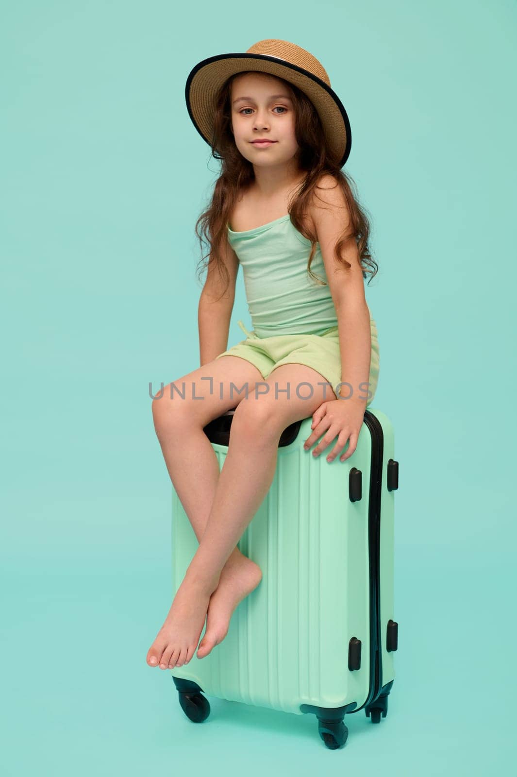 Emotional authentic little traveler girl, going for summer holidays, cutely smiles looking at camera, sitting on her polycarbonate trendy suitcase of turquoise color, isolated over blue background