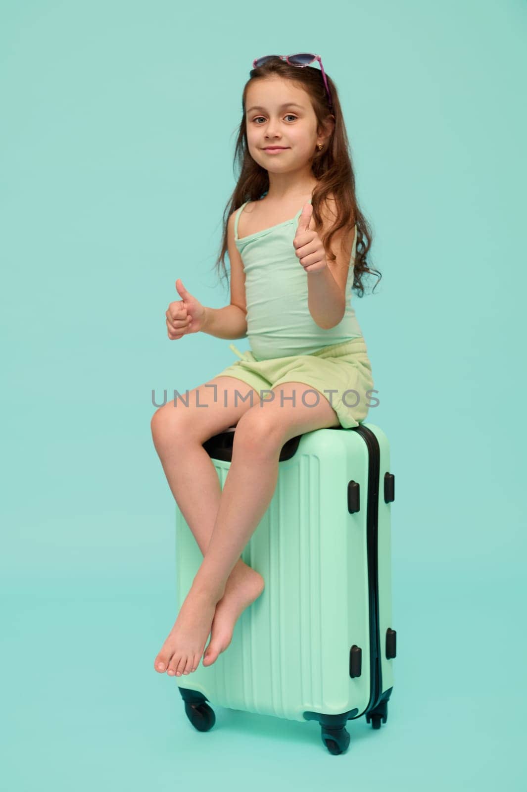 Emotional authentic little traveler girl, going for summer holidays, thumbs up, smiles looking at camera, sitting on her polycarbonate trendy suitcase of turquoise color, isolated over blue background