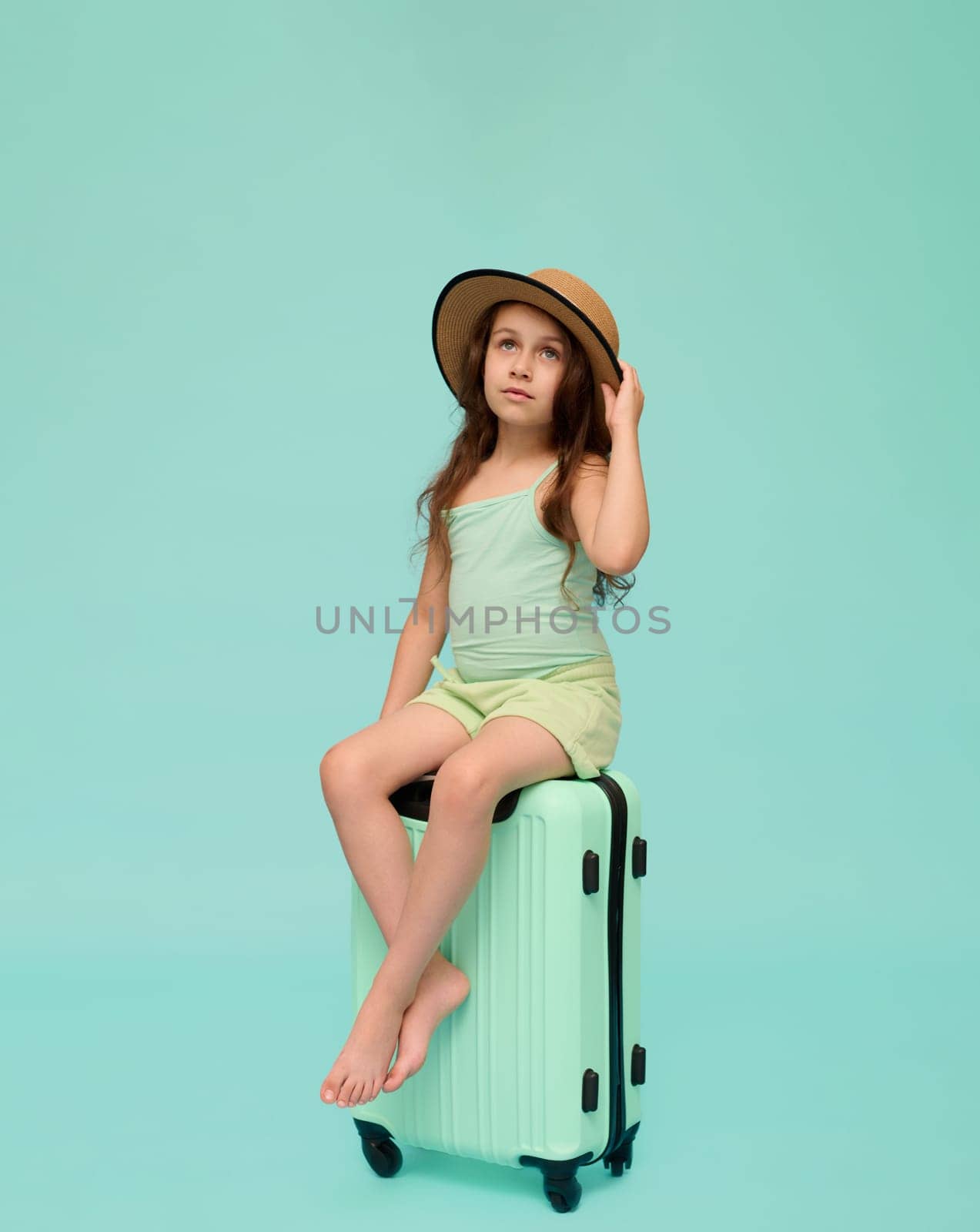 Vertical advertising portrait of a little child girl, passenger sitting on a trendy plastic valise, looking at camera, dressed in a straw hat and summer clothes, isolated over blue studio background