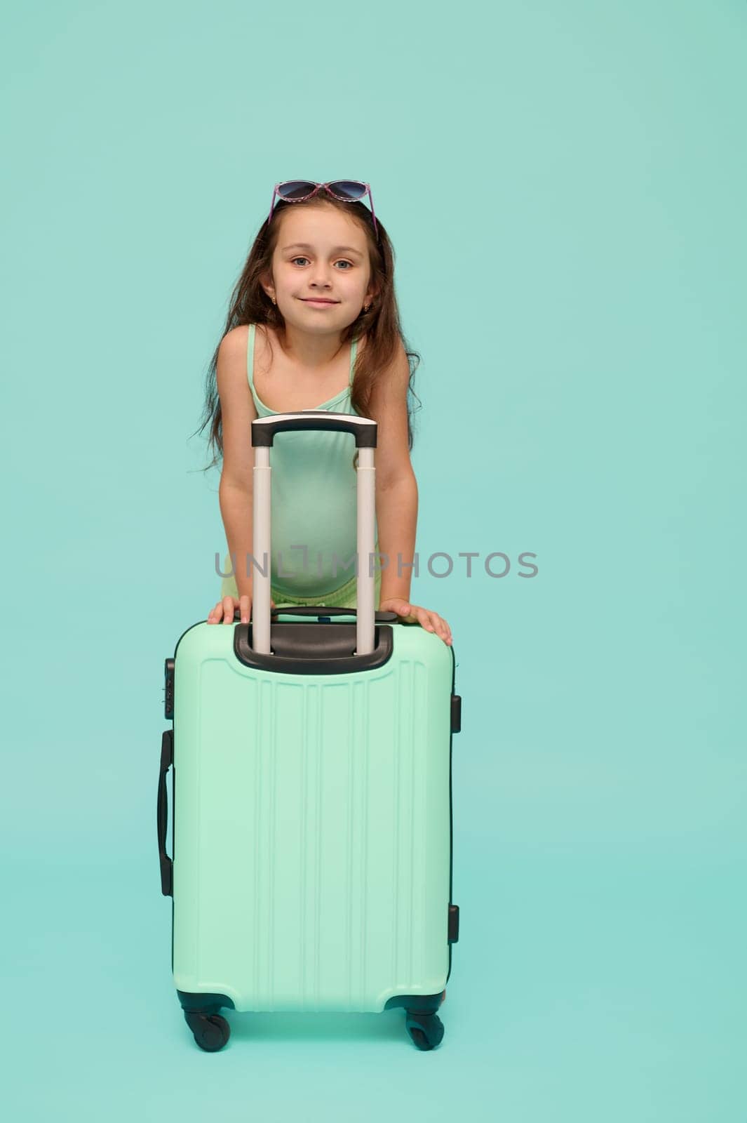 Caucasian happy child, beautiful little traveler girl with turquoise suitcase, smiles looking at camera, isolated on blue studio background. Kids. Travel. Weekend getaway and summer holidays concept