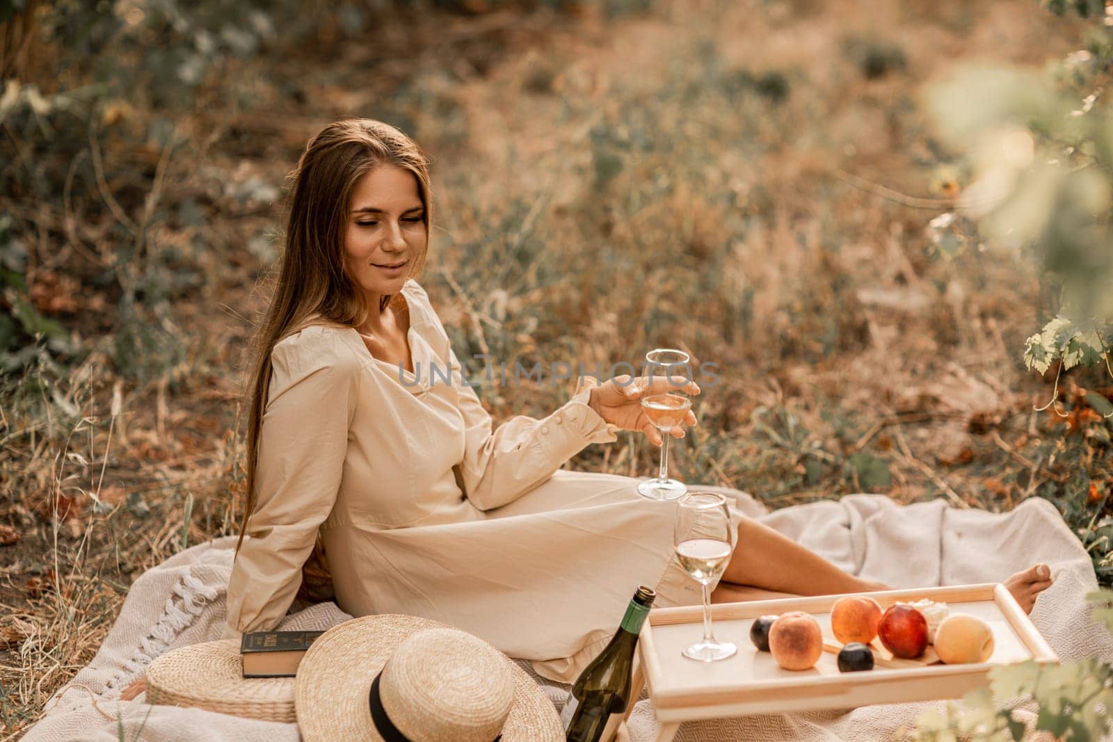 Woman picnic vineyard. Romantic dinner, fruit and wine. Happy woman with a glass of wine at a picnic in the vineyard on sunny day, wine tasting at sunset