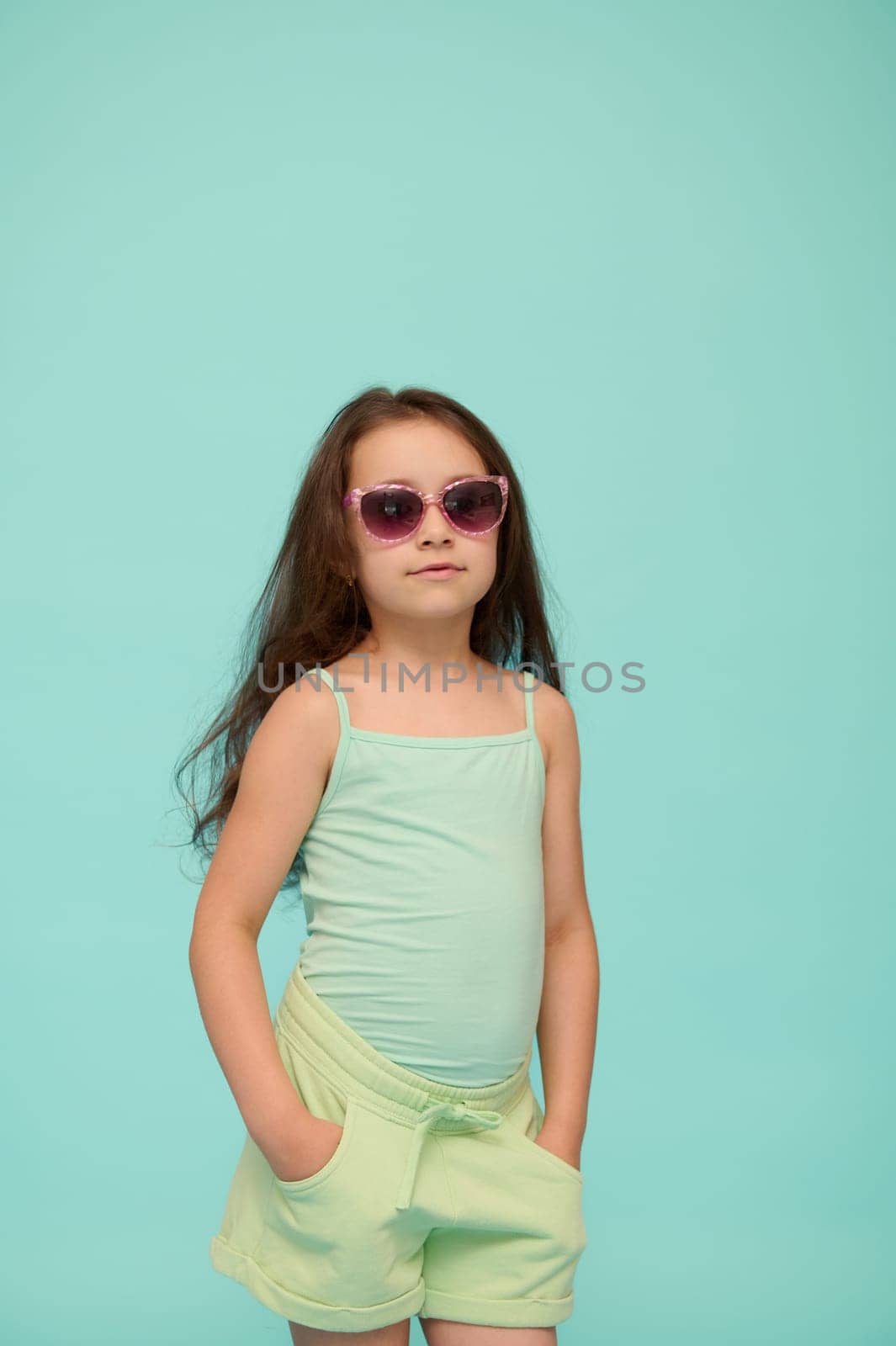 Adorable school age child girl in summer clothes and sunglasses, putting hands in pockets, confidently looking at camera, isolated on blue studio background with free advertising space for text